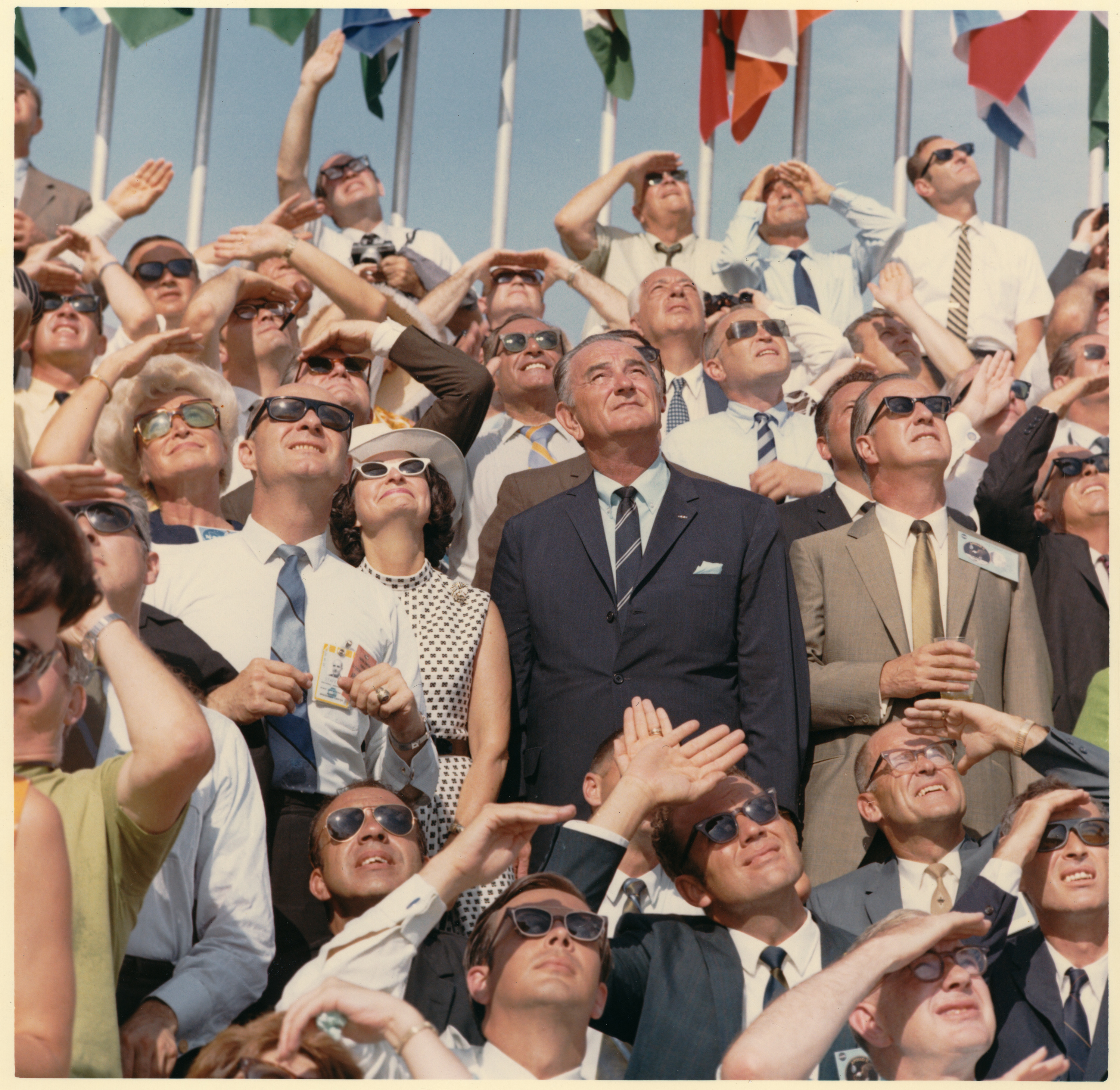 Lady Bird and Lyndon Johnson look to the sky amid a crowd of similar sky lookers, many with sunglasses.